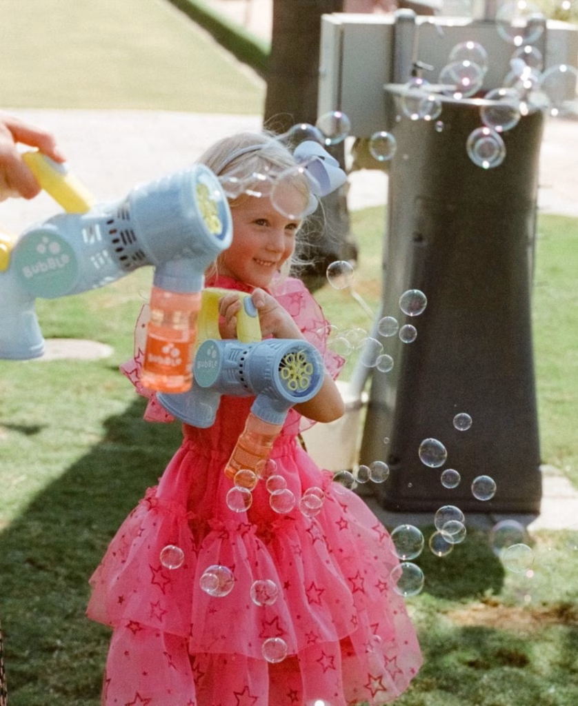 A whimsical color film image of a young flower girl blowing bubbles on the lawn, captured with soft-focus greenery, warm nostalgic tones, and organic grain by Phoebe Kate Photography.
