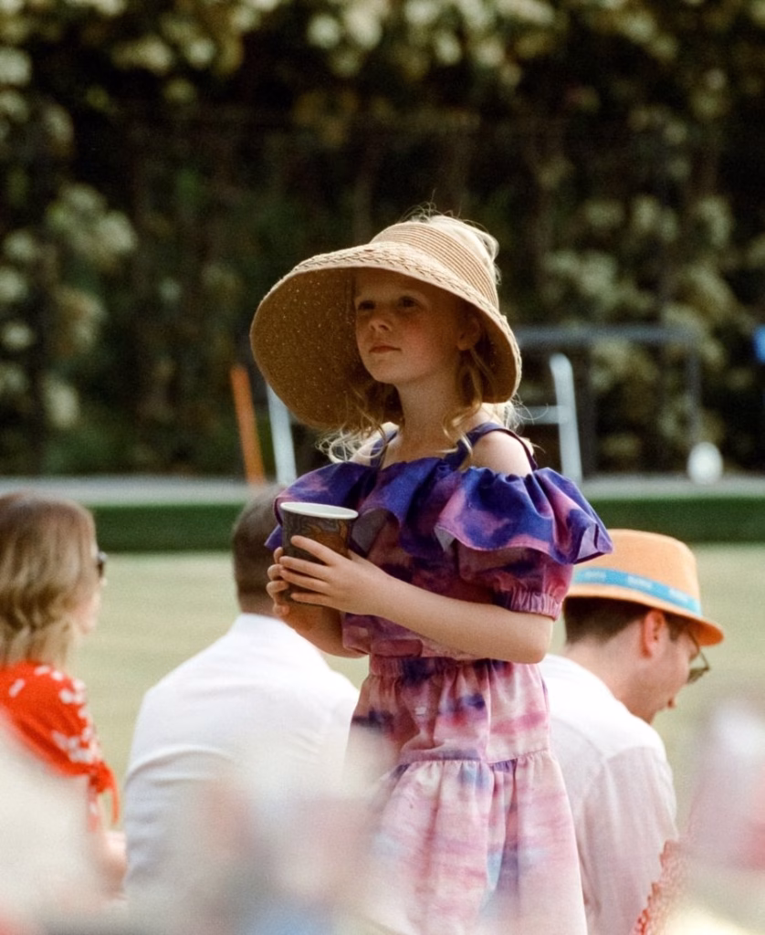 A candid, sun-drenched color film portrait of a flower girl in a detailed lace outfit, captured during a quiet moment at a wedding with a painterly, nostalgic aesthetic by Phoebe Kate Photography.