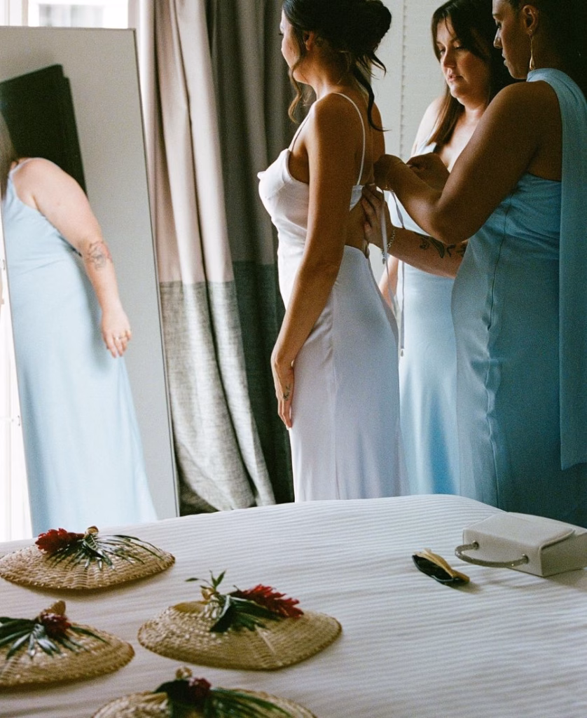 A documentary-style film photograph of a bridesmaid helping the bride into her gown, capturing the quiet anticipation of the wedding morning.