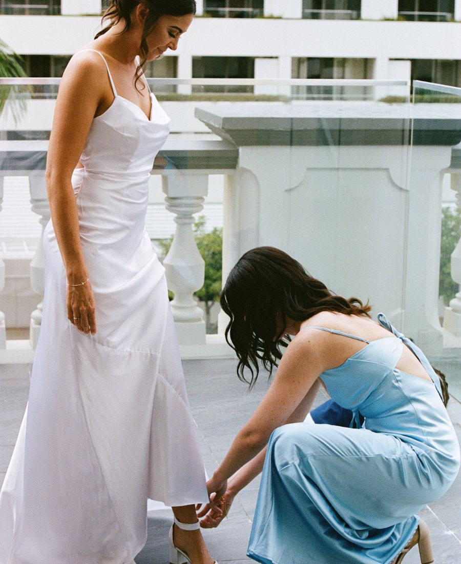 A candid, color 35mm film photograph of a bridesmaid helping a bride with her wedding shoes. Captured with a nostalgic editorial aesthetic, featuring natural morning light, organic film grain, and soft pastel tones in a Cairns wedding setting.