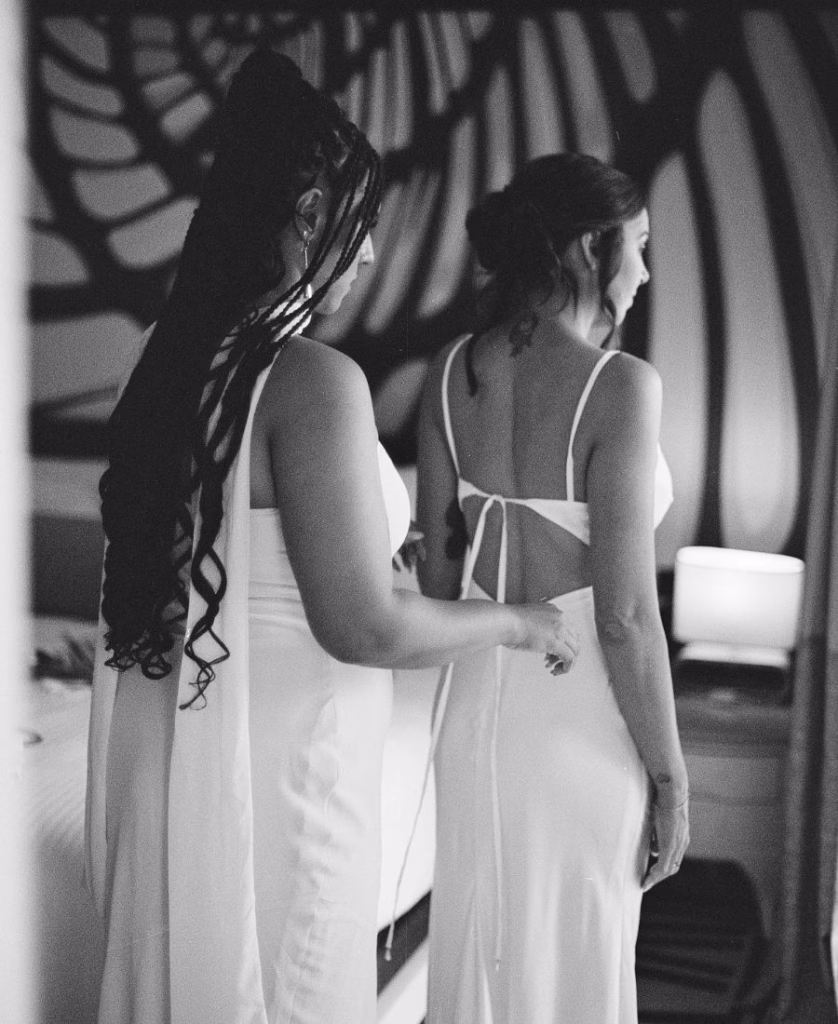 A black and white close-up film shot of a bridesmaid’s hands tying the delicate lace of the bride’s dress, emphasizing texture and connection.